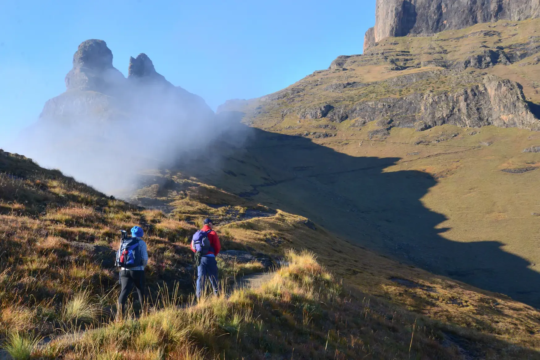 Zwei Trekker auf einem schmalen Pfad in den südafrikanischen Drakensbergen blicken auf markante, nebelverhangene Felsgipfel.