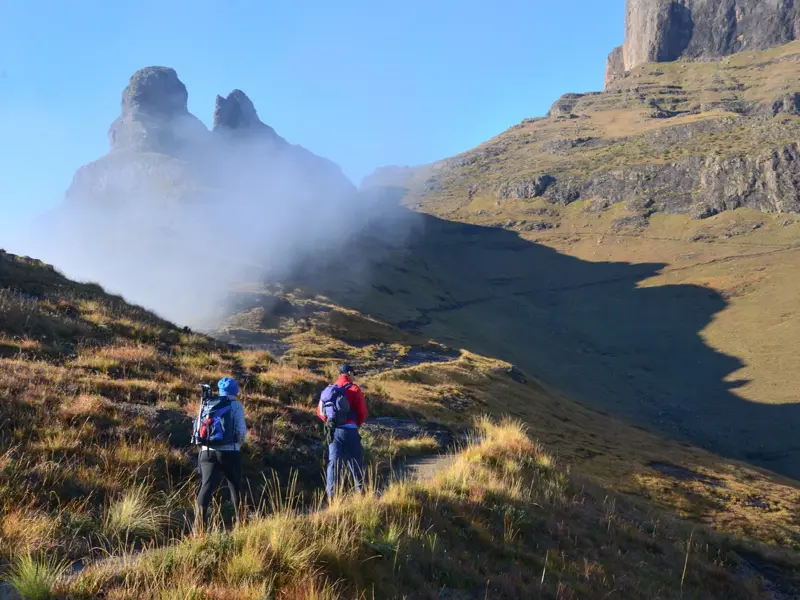 Zwei Trekker auf einem schmalen Pfad in den südafrikanischen Drakensbergen blicken auf markante, nebelverhangene Felsgipfel.