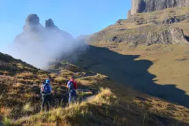 Zwei Trekker auf einem schmalen Pfad in den südafrikanischen Drakensbergen blicken auf markante, nebelverhangene Felsgipfel.