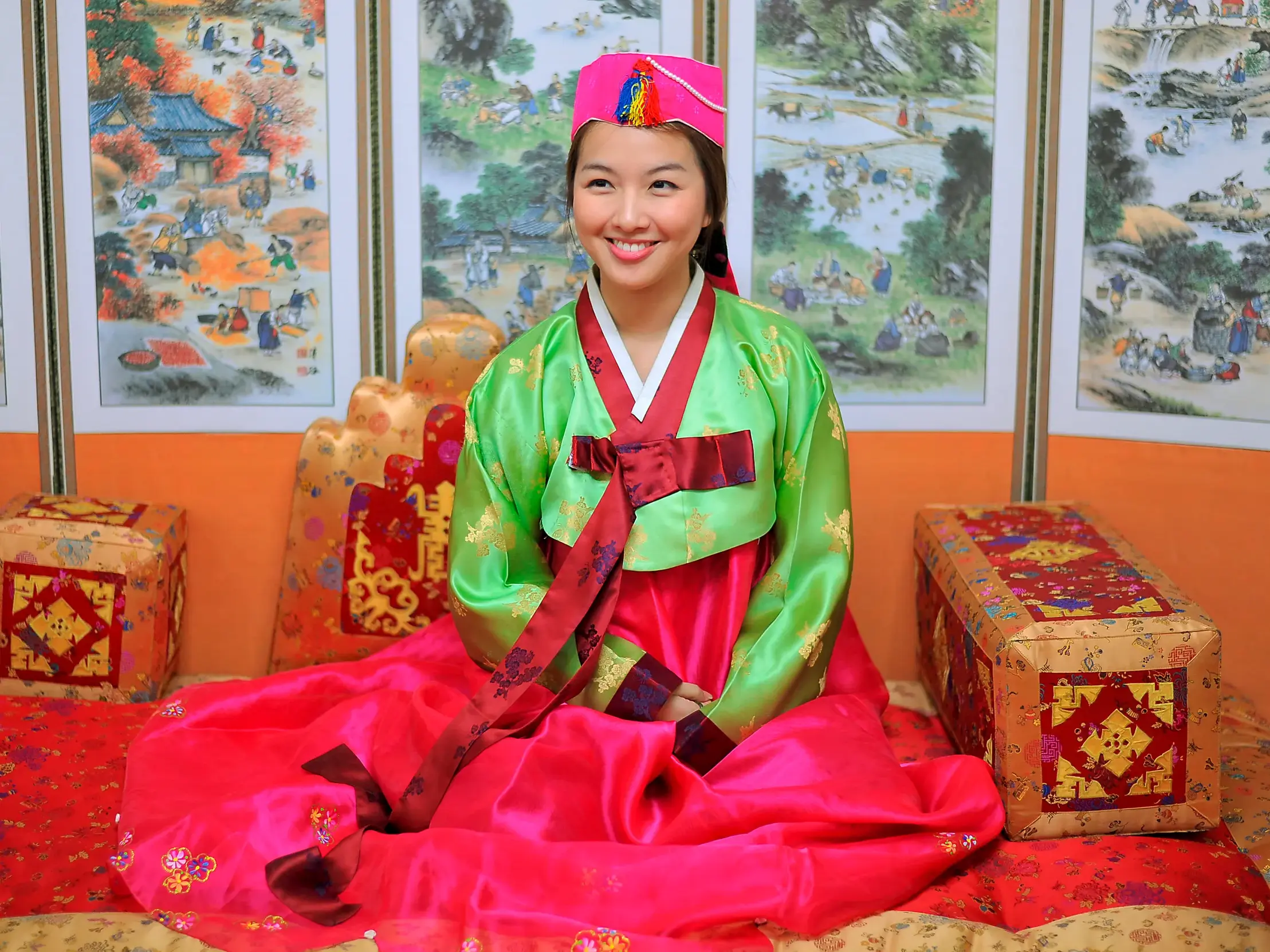 Eine lächelnde Frau in einem farbenfrohen, traditionellen Hanbok posiert sitzend in Südkorea vor einer bemalten Faltwand.