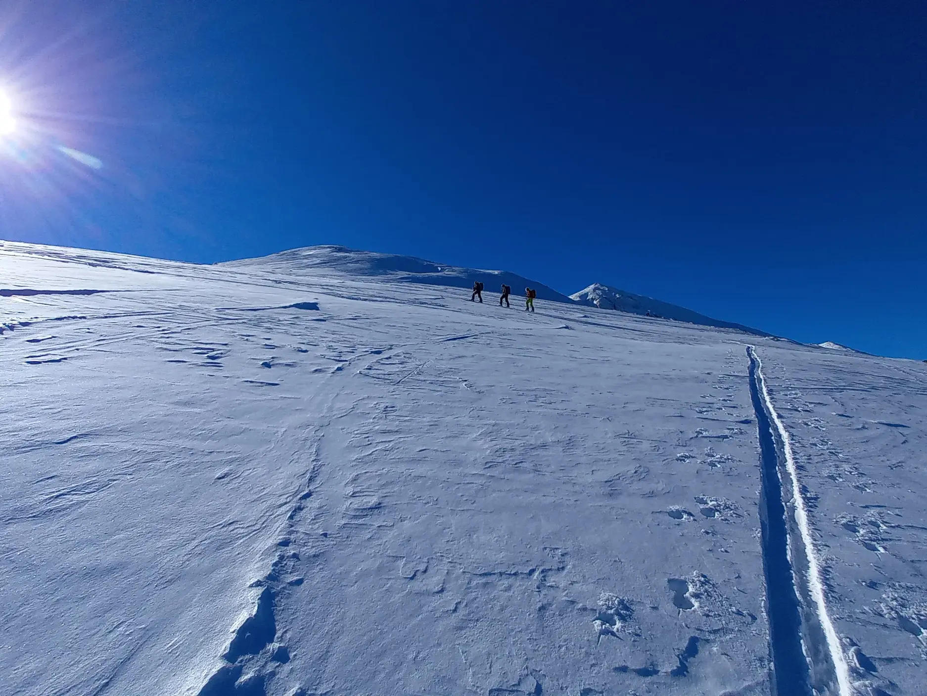 Drei Skitourengeher folgen einer Spur durch tiefen Schnee auf einen Gipfel im Korab-Gebirge in Albanien bei Sonnenschein.