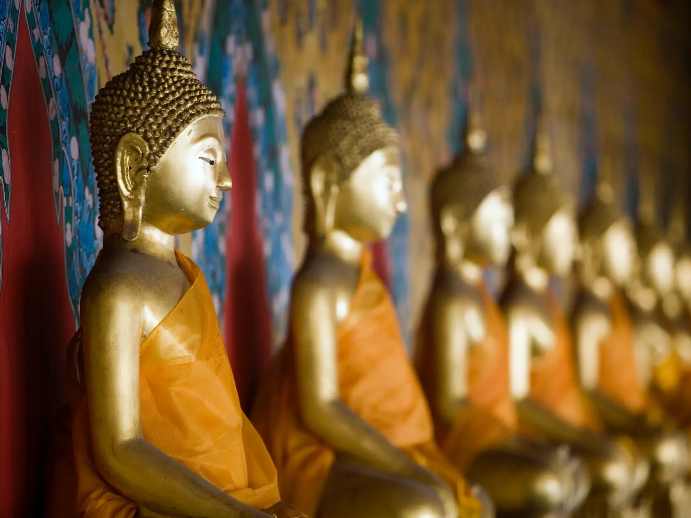 Goldene Buddha-Statue im Profil, aufgereiht mit weiteren Figuren in orangefarbenen Roben in einem Tempel in Thailand.