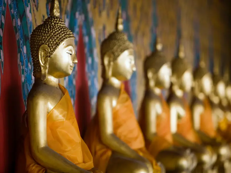 Goldene Buddha-Statue im Profil, aufgereiht mit weiteren Figuren in orangefarbenen Roben in einem Tempel in Thailand.