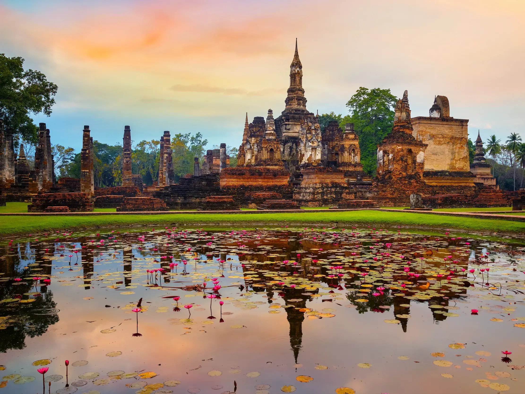Die Ruinen des Wat Mahathat im Geschichtspark Sukhothai spiegeln sich bei Sonnenuntergang im von Lotusblüten bedeckten See.