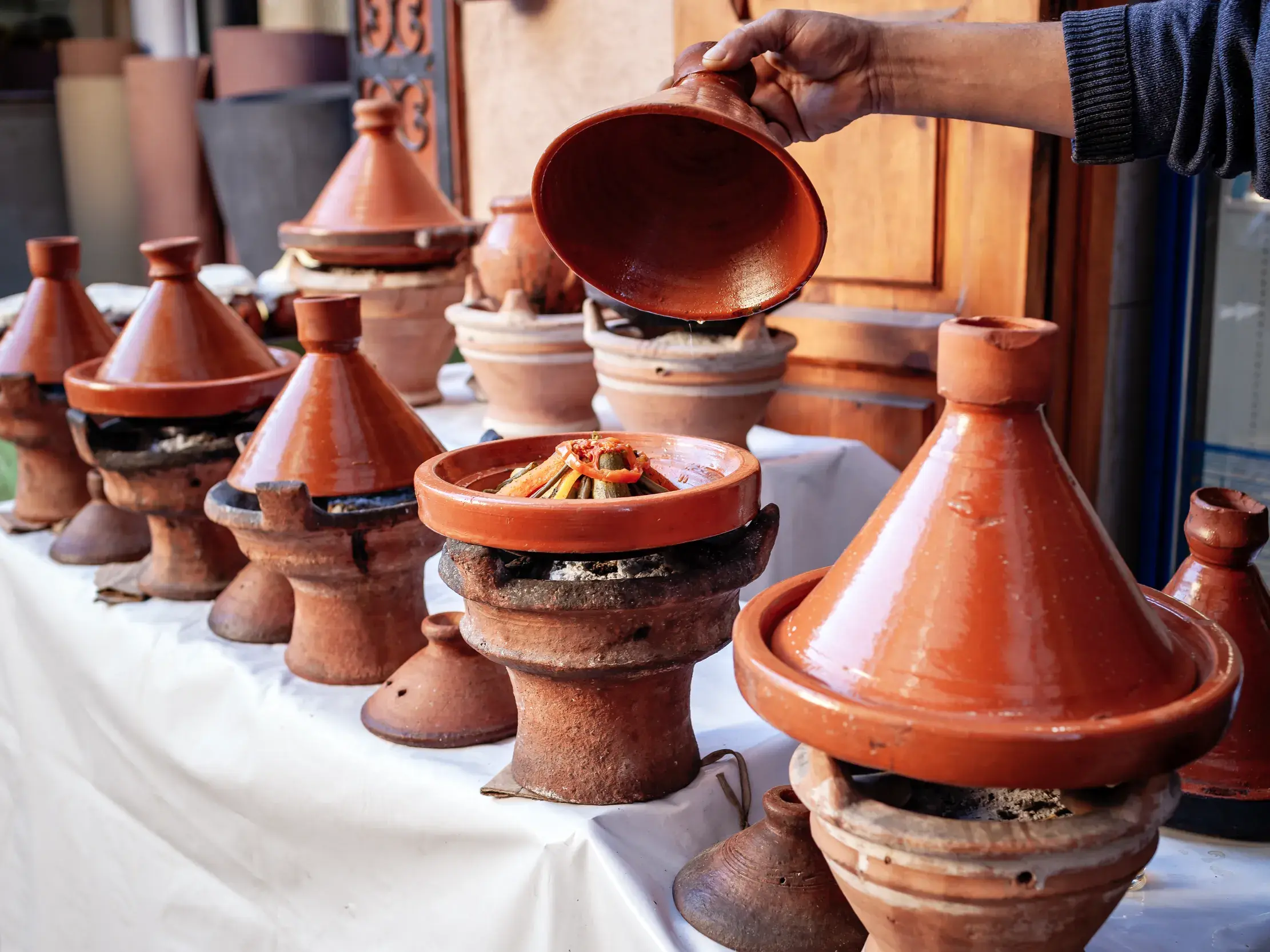 Eine Person hebt den Deckel eines traditionellen Tajine-Tontopfs und enthüllt ein dampfendes Gericht bei einem Kochkurs in Marokko.