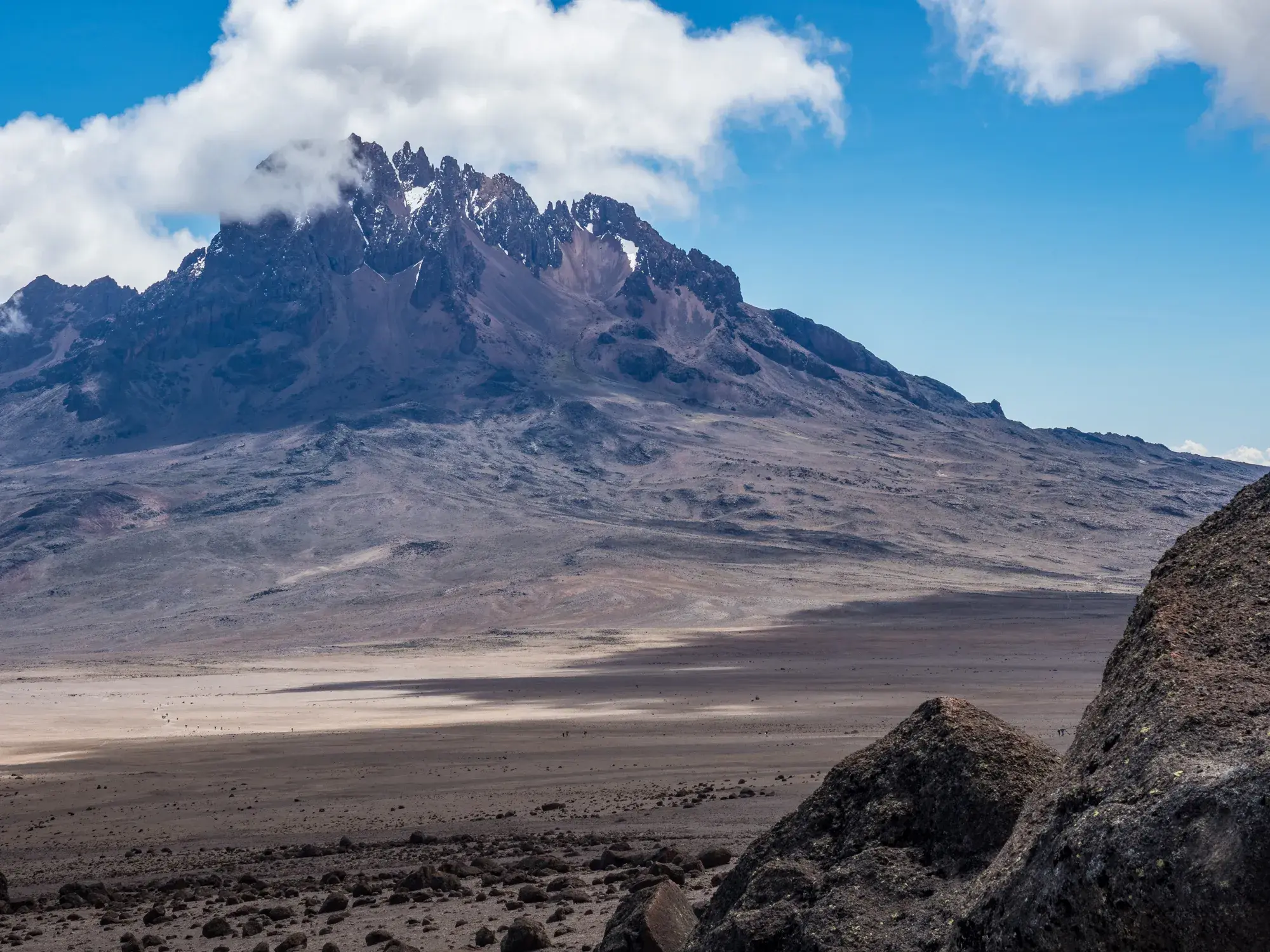 Weite, karge Hochebene am Kilimanjaro vor dem felsigen Mawenzi-Gipfel, dessen Spitzen von Wolken umspielt werden.