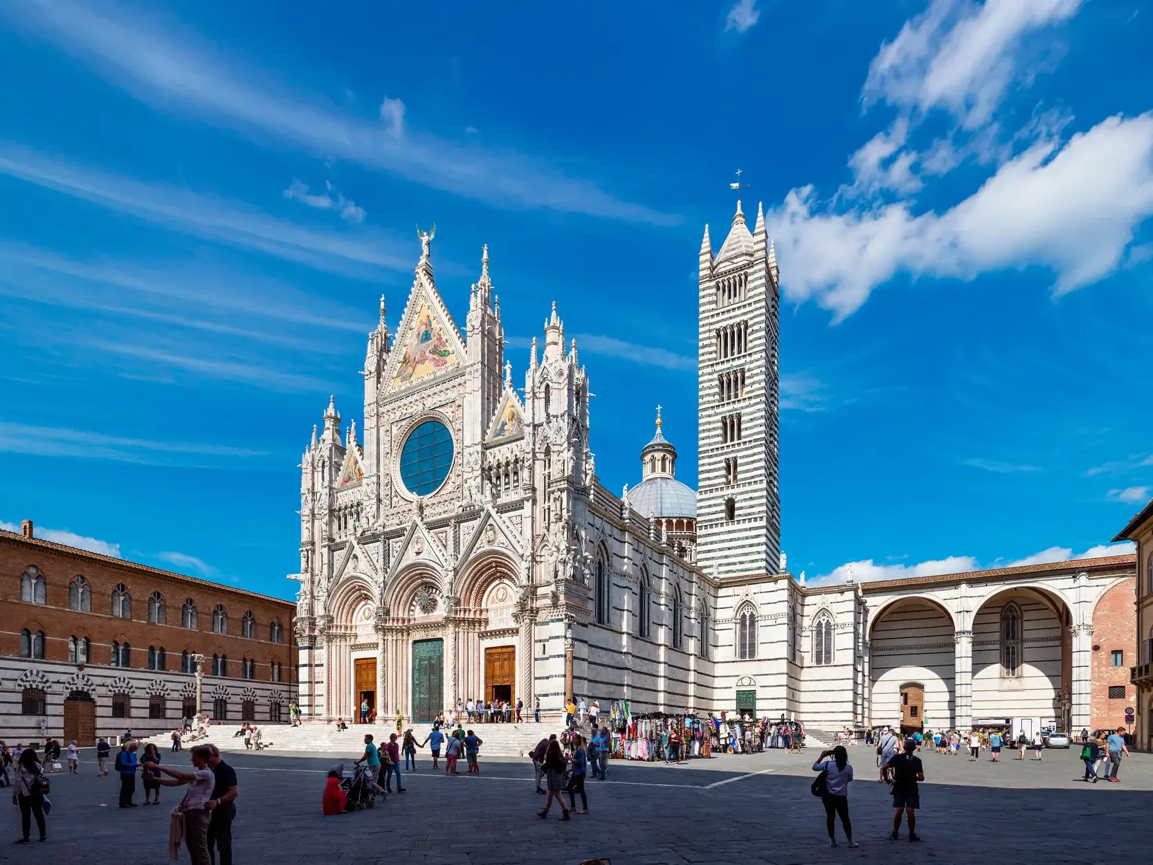 Majestätische Fassade des zebragestreiften Doms von Siena bei Sonnenschein, davor belebter Platz mit zahlreichen Besuchern.