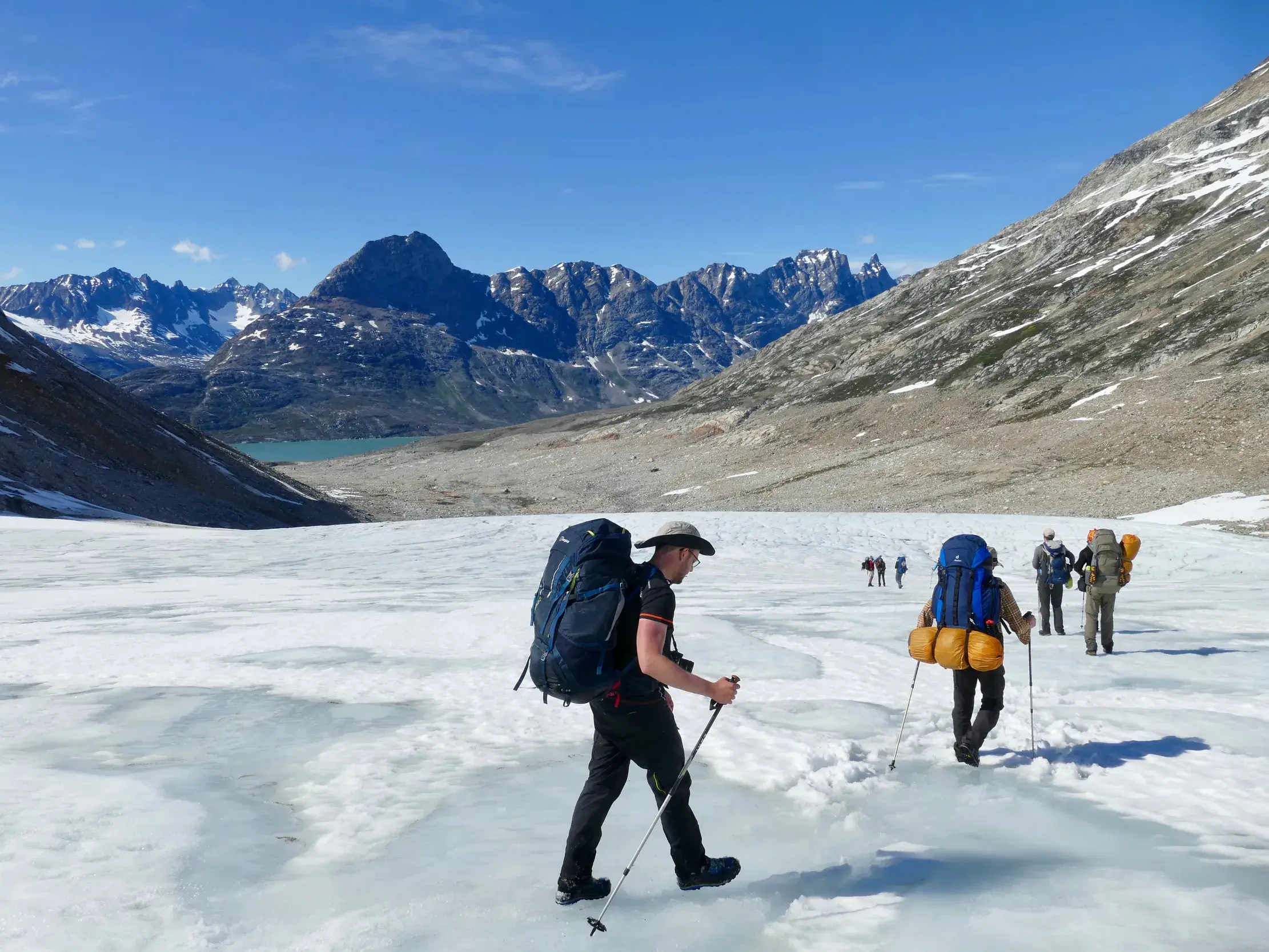 Wanderer mit Trekkingstöcken und großen Rucksäcken überqueren einen Gletscher in der einsamen Wildnis Ostgrönlands.