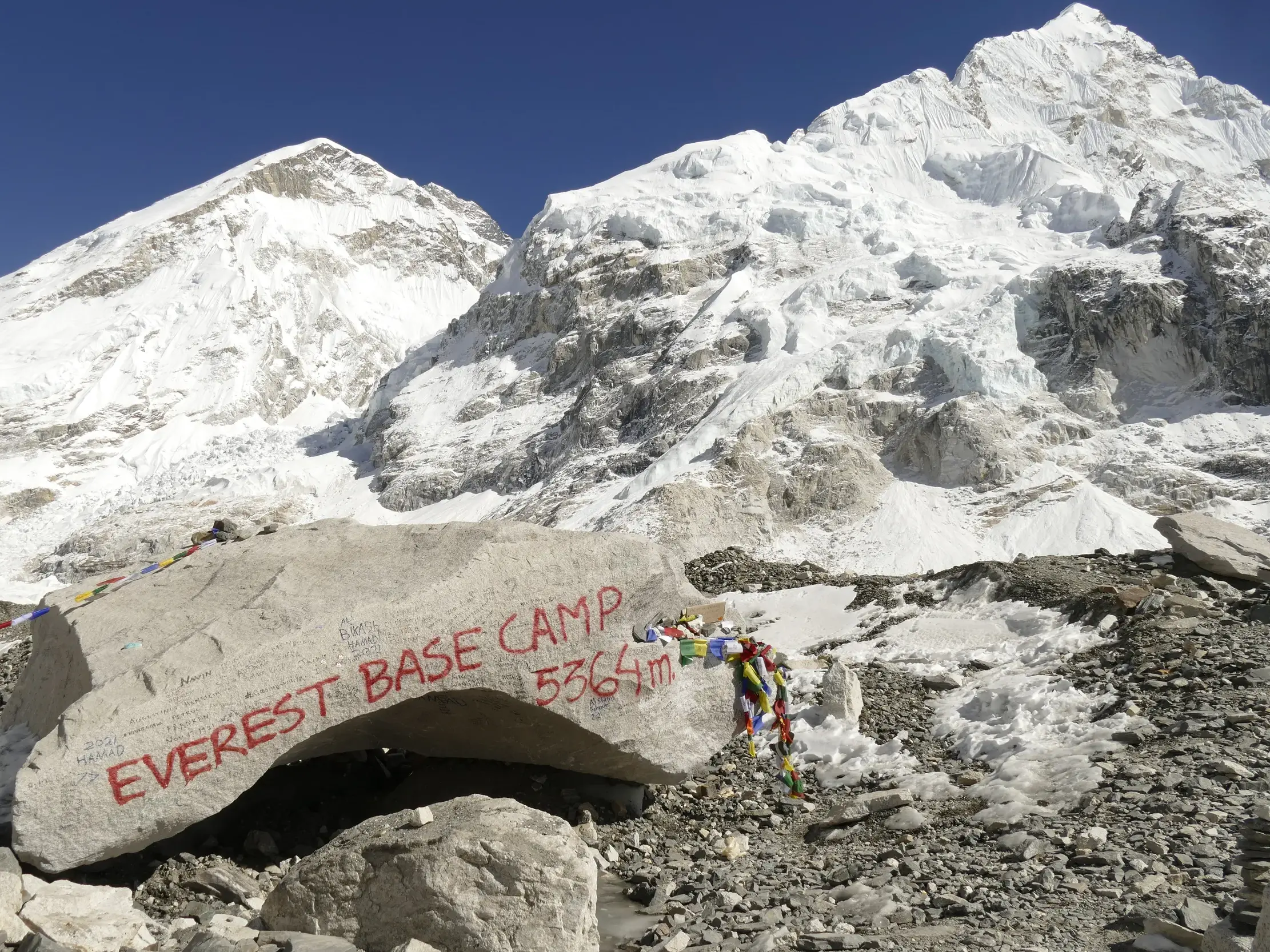 Der beschriftete Fels des Everest-Basislagers mit Gebetsfahnen vor der schneebedeckten Bergkulisse des Khumbu-Himalaya.