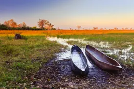 Zwei traditionelle Einbaum-Boote (Mokoros) am Ufer der weiten Graslandschaft des Okavango Deltas bei Sonnenuntergang.