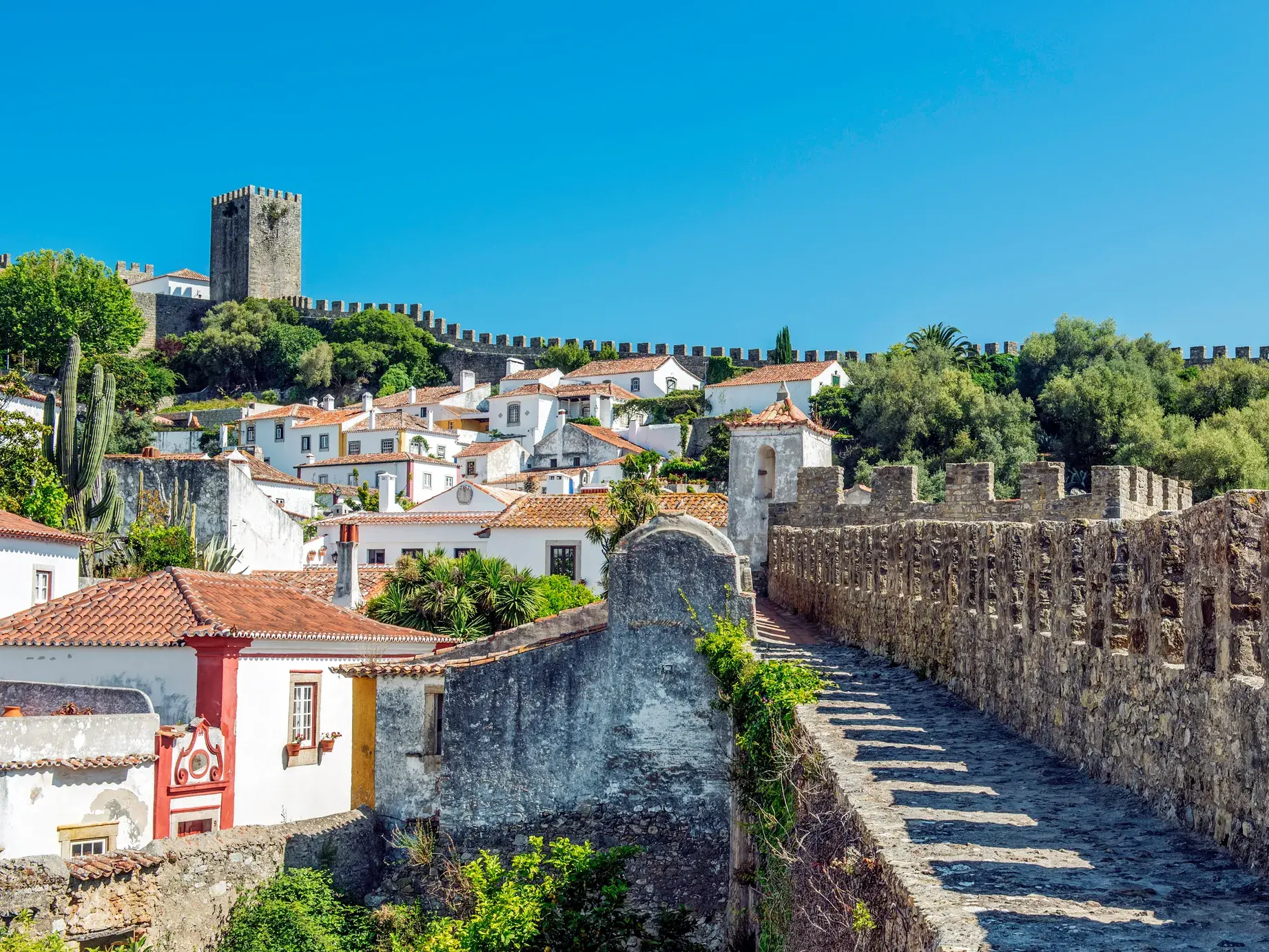 Blick vom begehbaren, mittelalterlichen Mauergürtel auf die weißen Häuser des Bilderbuchdorfs Obidos bei strahlend blauem Himmel.
