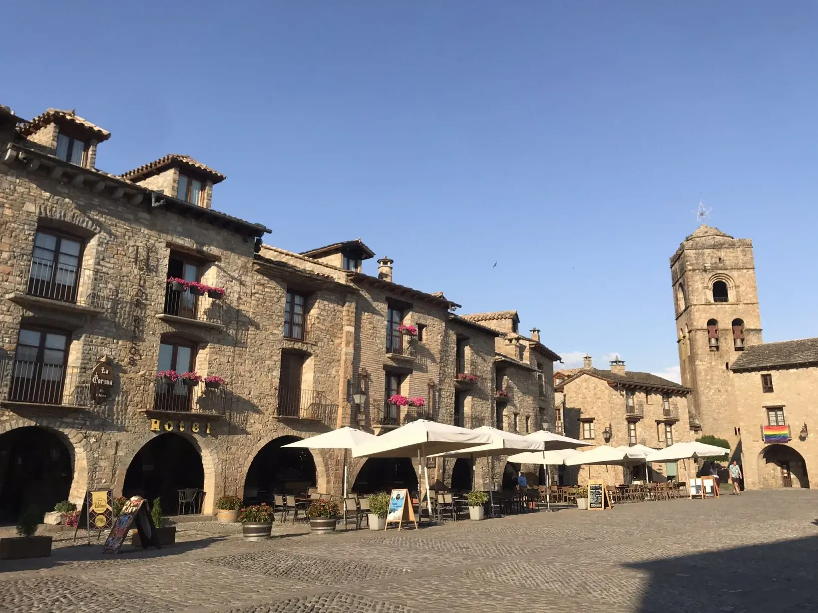 Sonniger Dorfplatz in Alquézar in Aragonien mit historischen Steinhäusern, Cafés und einem Glockenturm im Hintergrund.