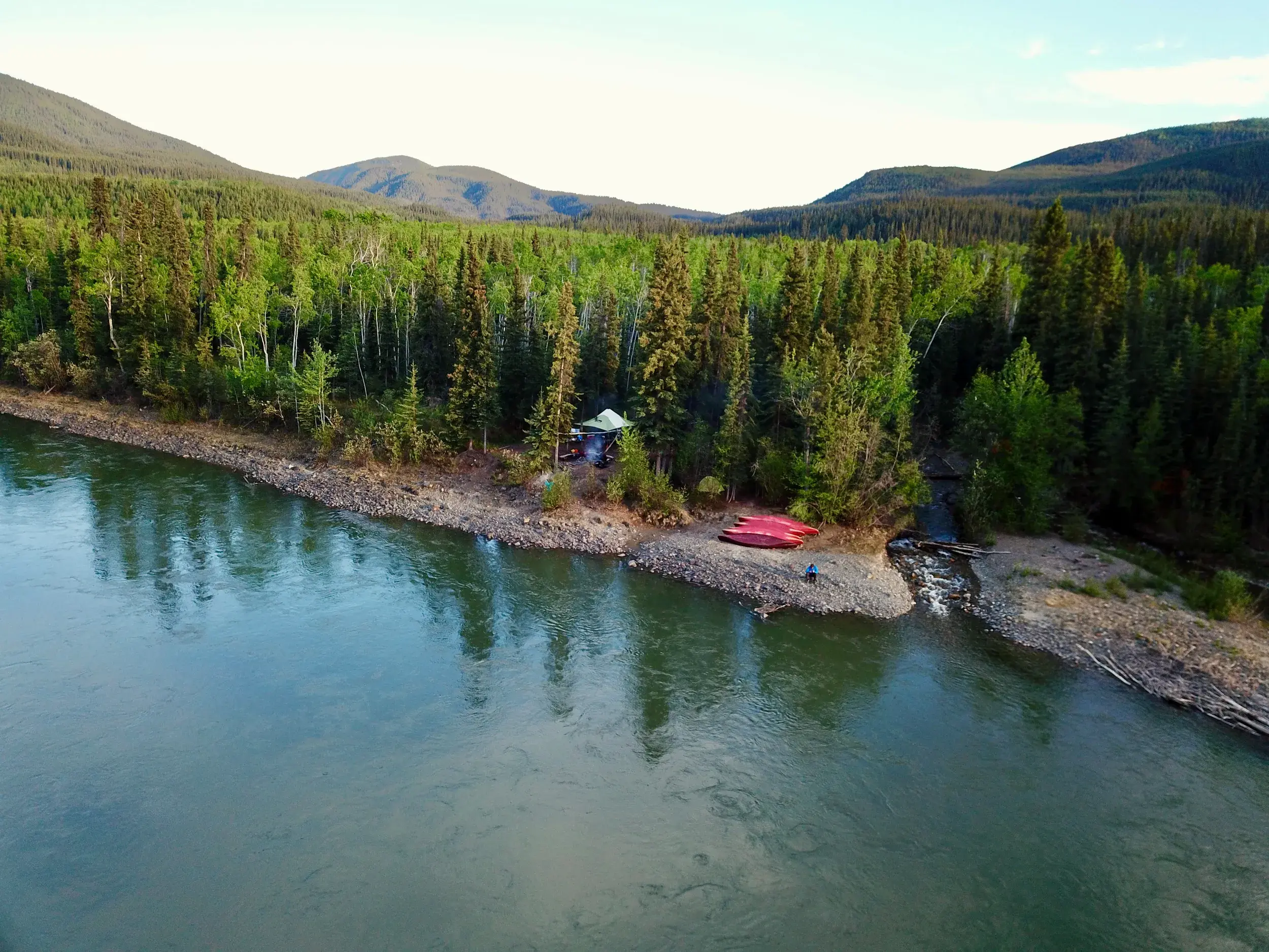 Zeltlager mit zwei roten Kanus am Ufer des Yukon River in Kanadas Wildnis, im Hintergrund dichte Wälder und Berge.