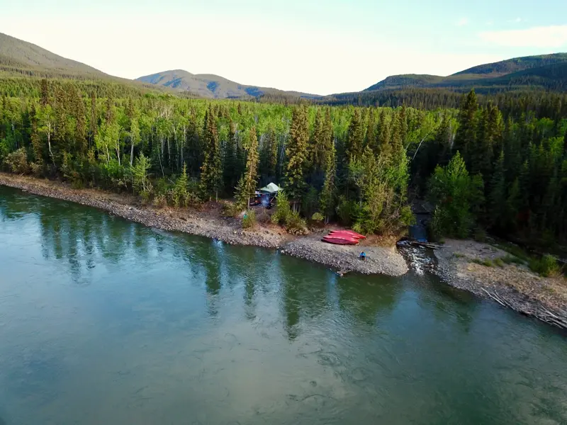 Zeltlager mit zwei roten Kanus am Ufer des Yukon River in Kanadas Wildnis, im Hintergrund dichte Wälder und Berge.