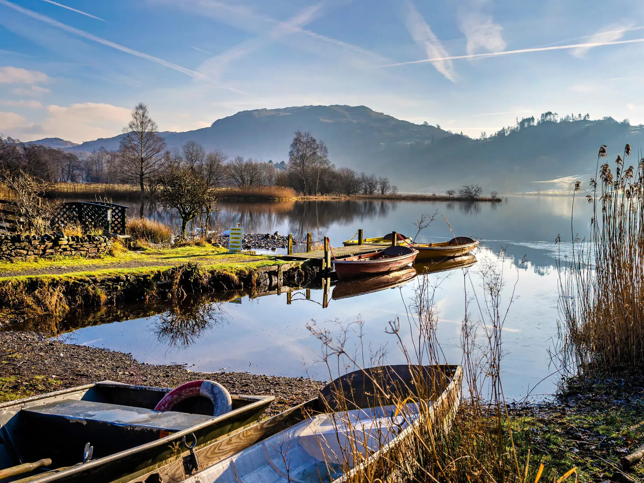 Ruderboote an einem Steg am Ufer eines stillen Sees im englischen Lake District, dessen Berglandschaft sich im Wasser spiegelt.