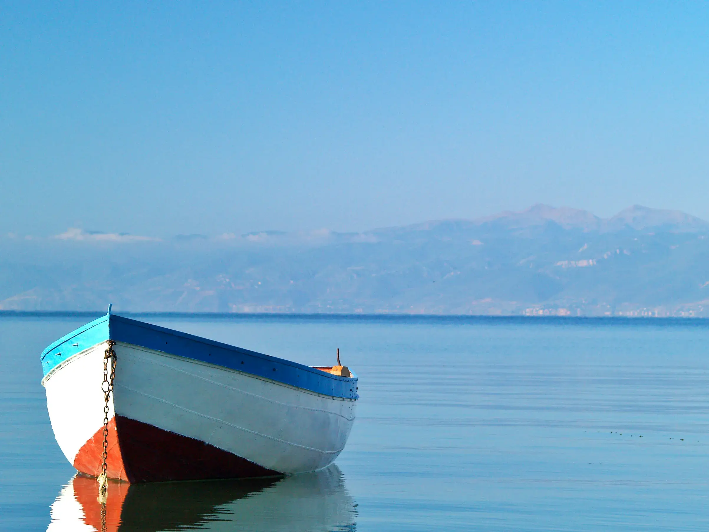 Ein weiß-blau-rotes Fischerboot ankert im ruhigen, blauen Wasser des Ohrid-Sees in Nordmazedonien bei strahlendem Sonnenschein.