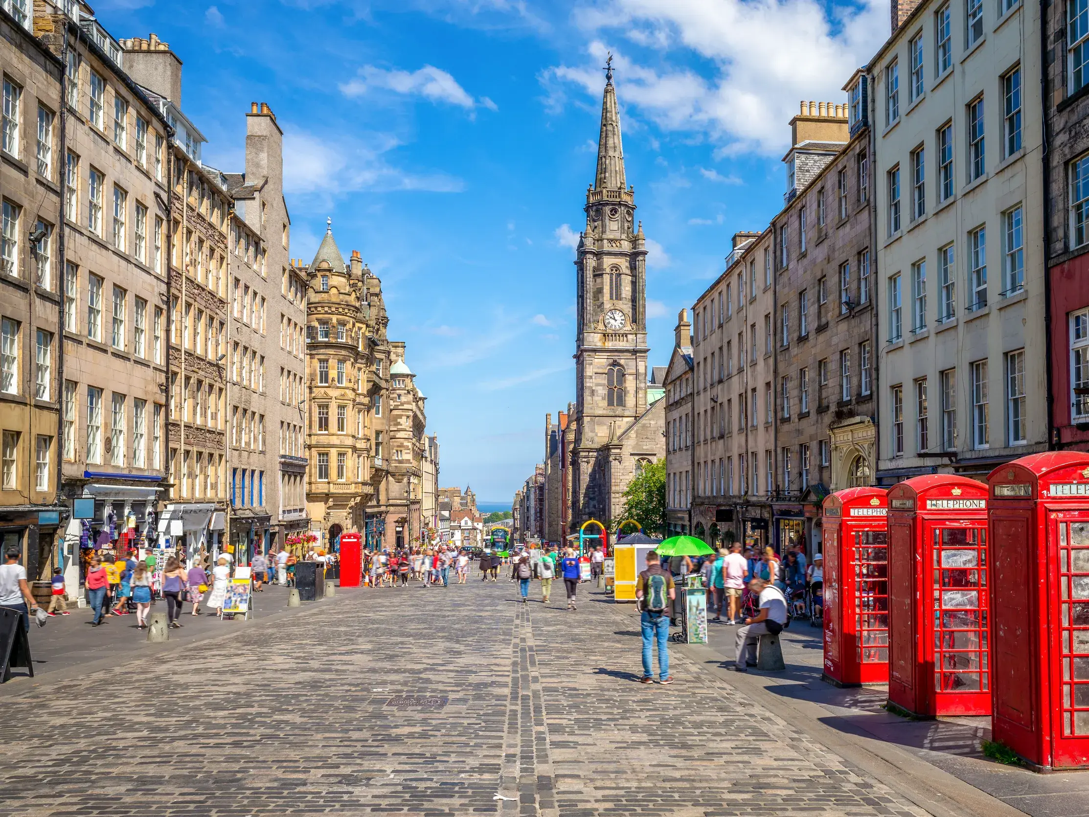 Die Royal Mile in Edinburgh mit historischen Gebäuden, dem Turm der Tron Kirk und roten Telefonzellen an einem sonnigen Tag.