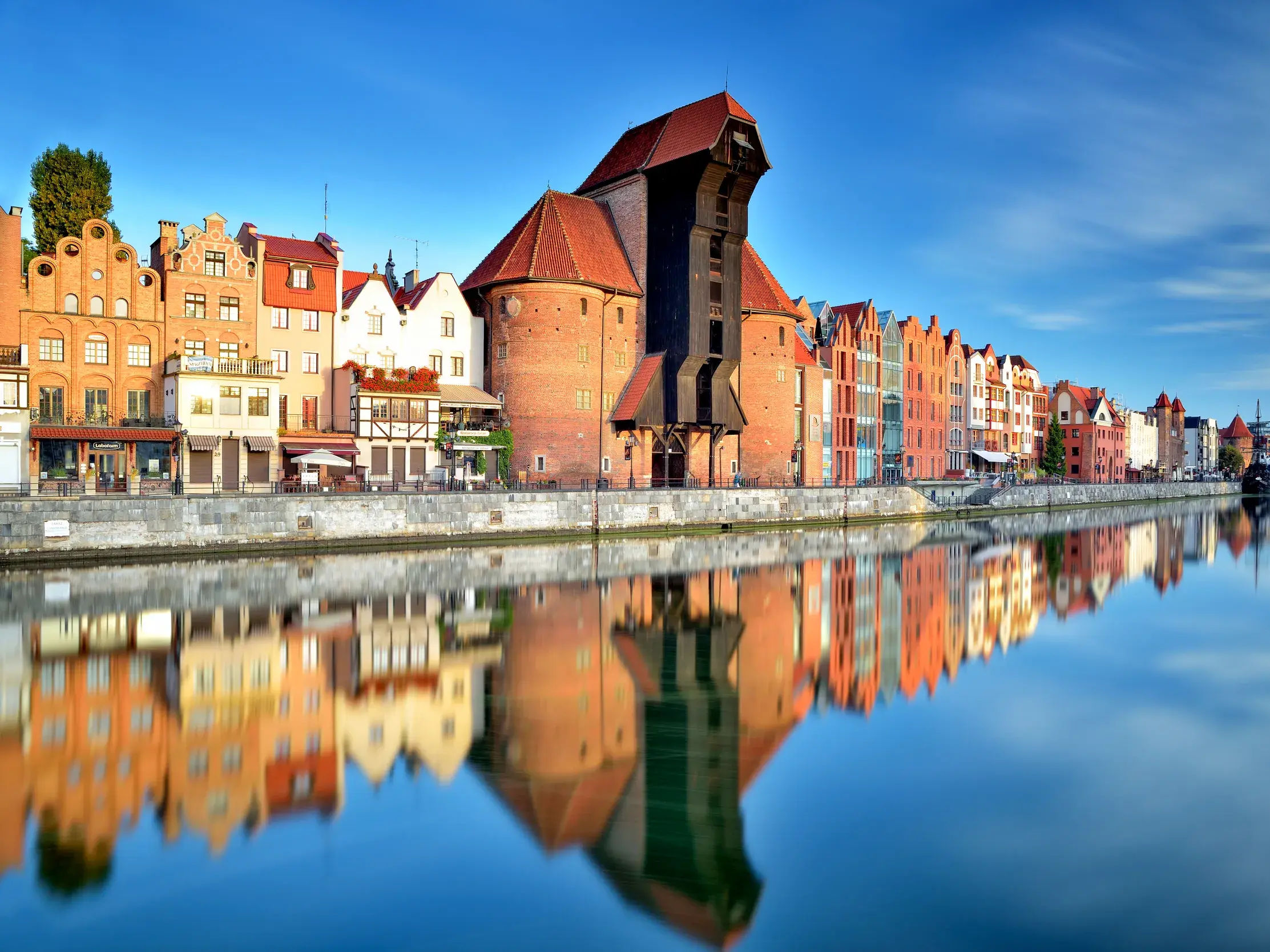 Historische Patrizierhäuser und das berühmte Krantor in Danzig spiegeln sich an einem sonnigen Tag im ruhigen Wasser.