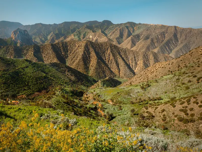 Panoramablick beim Wandern auf La Gomera über grüne Terrassenfelder auf die zerklüftete Berglandschaft mit dem Roque de Agando.