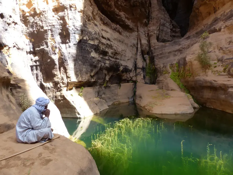 Ein Tuareg-Reisebegleiter in blauer Tracht ruht an einem smaragdgrünen Wasserloch in einem Canyon in der algerischen Sahara.
