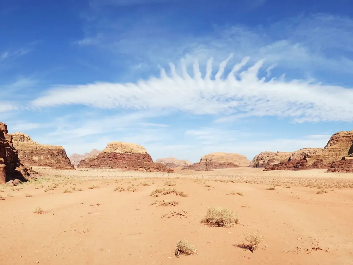 Weite Sandlandschaft der Wadi Rum Wüste in Jordanien mit roten Felsen und einer federartigen Wolke am blauen Himmel.