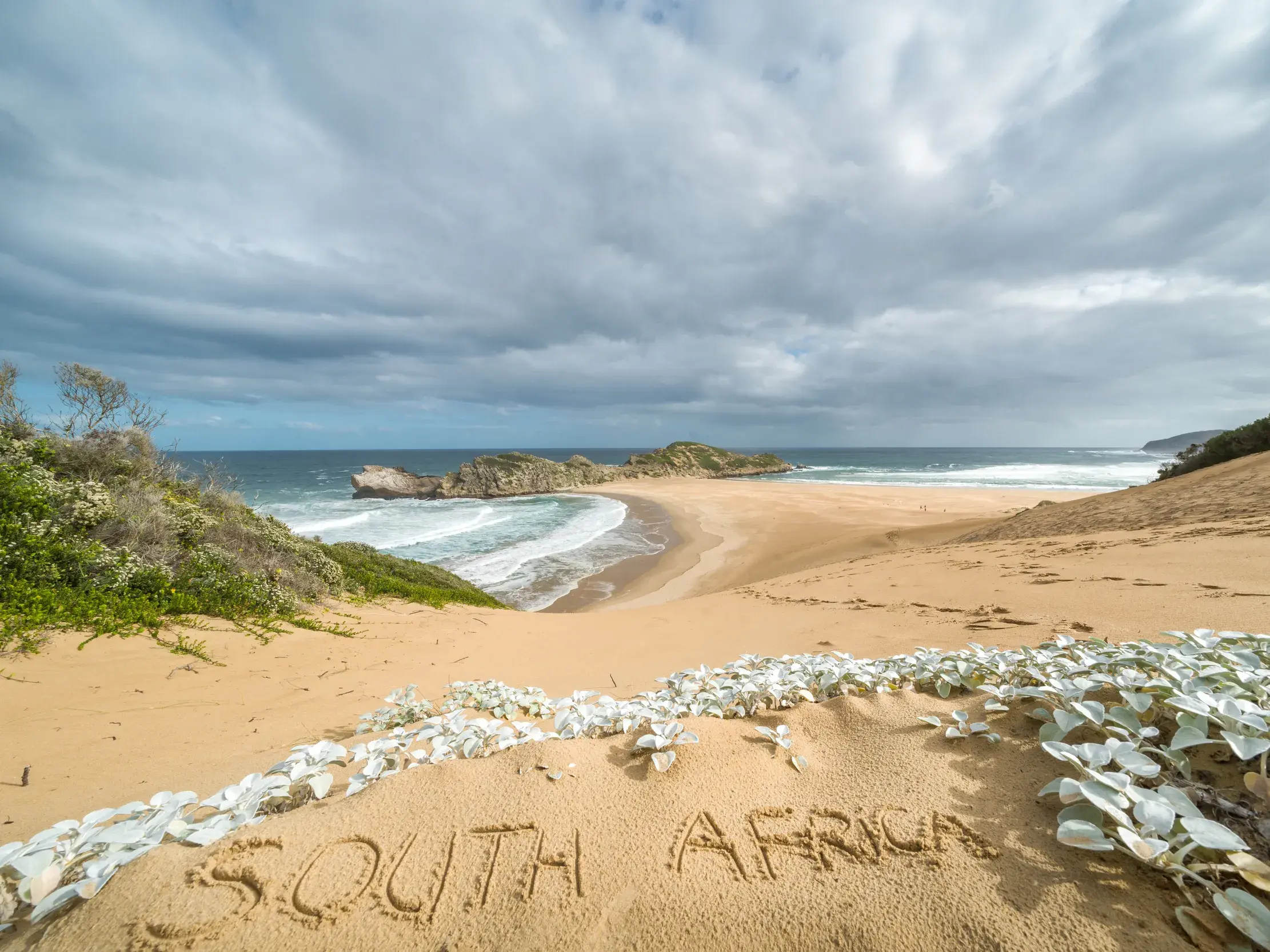Schriftzug "South Africa" im Sand einer Düne mit Blick auf die wilde Küste der Garden Route unter wolkigem Himmel.