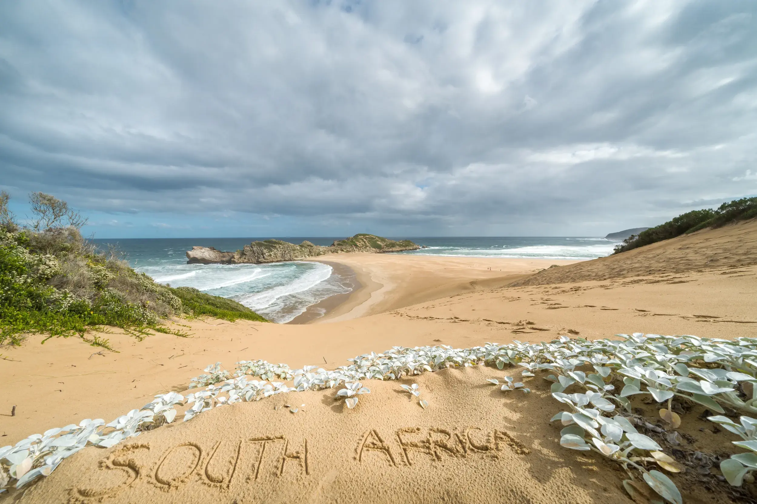 Schriftzug "South Africa" im Sand einer Düne mit Blick auf die wilde Küste der Garden Route unter wolkigem Himmel.