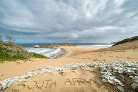 Schriftzug "South Africa" im Sand einer Düne mit Blick auf die wilde Küste der Garden Route unter wolkigem Himmel.