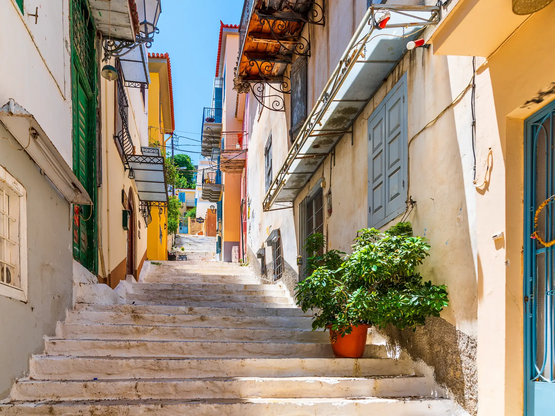 Eine steile Steintreppe führt bergauf durch eine enge Gasse in Nauplio, gesäumt von bunten Häusern und Balkonen unter blauem Himmel.
