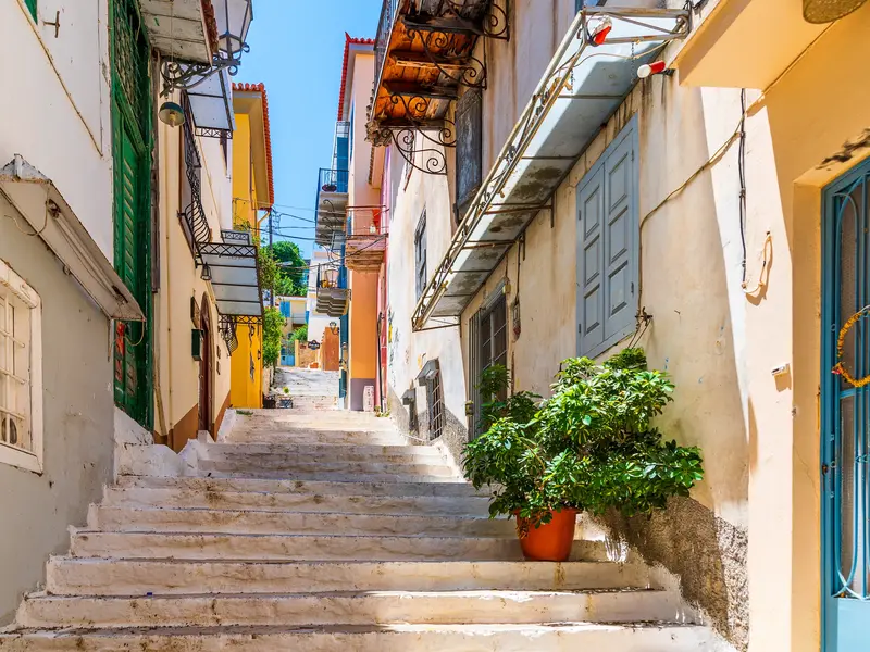 Eine steile Steintreppe führt bergauf durch eine enge Gasse in Nauplio, gesäumt von bunten Häusern und Balkonen unter blauem Himmel.