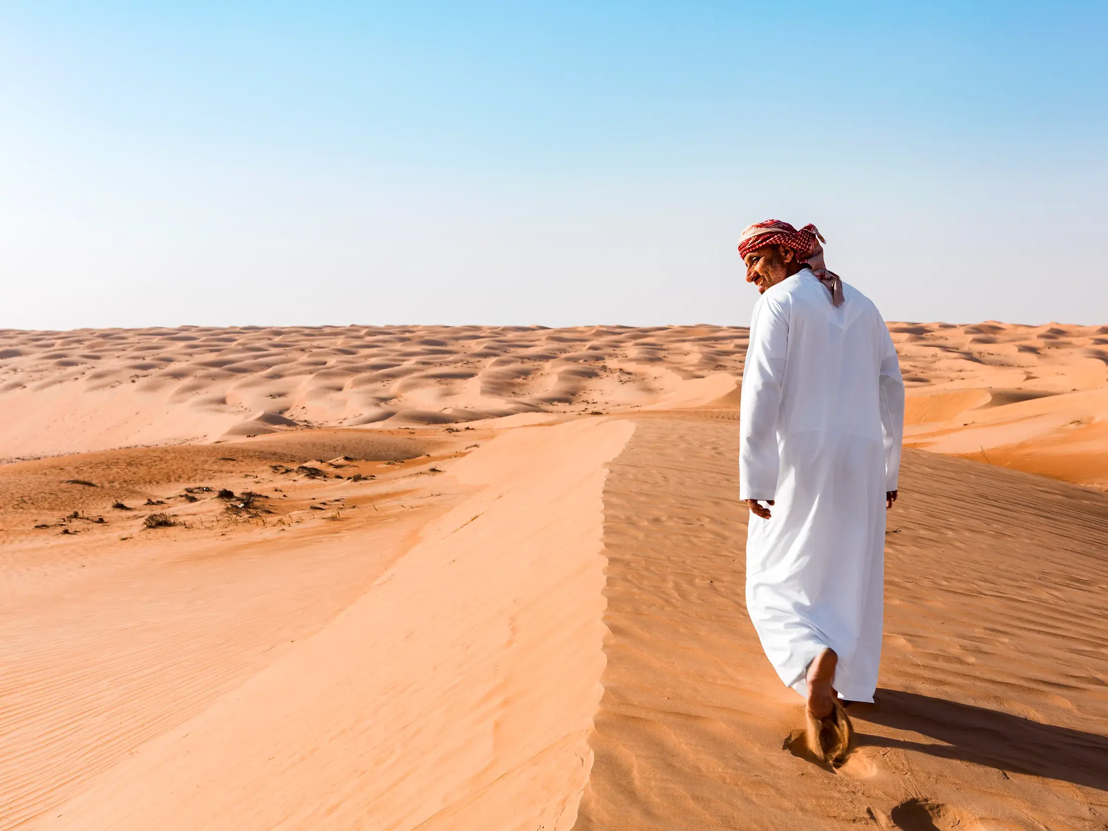 Mann in traditioneller omanischer Kleidung wandert über eine Sanddüne in der weiten Wüstenlandschaft der Wahiba Sands im Oman.