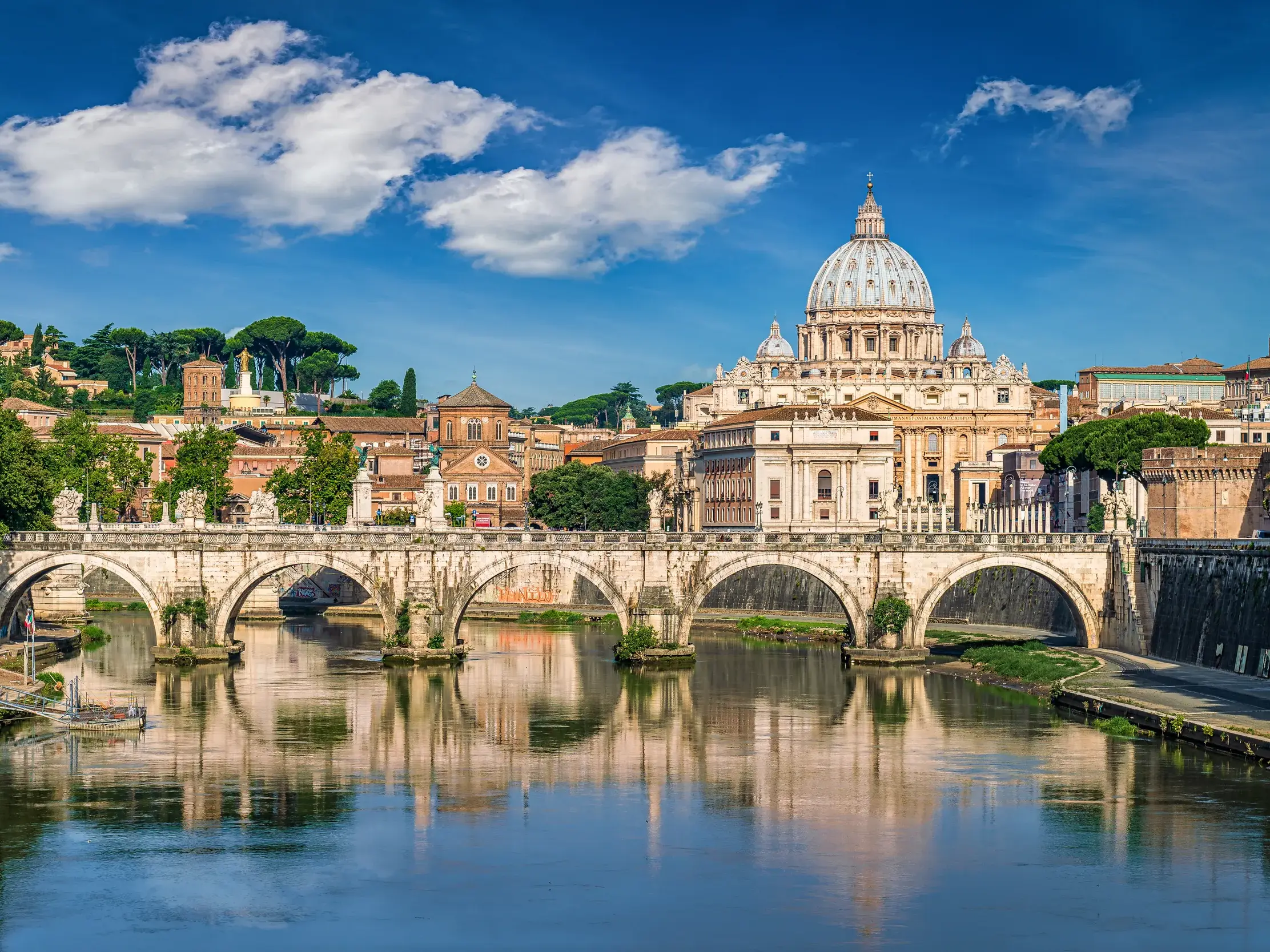 Sonniger Blick über den Tiber auf die majestätische Peterskirche in Rom mit einer historischen Bogenbrücke im Vordergrund.