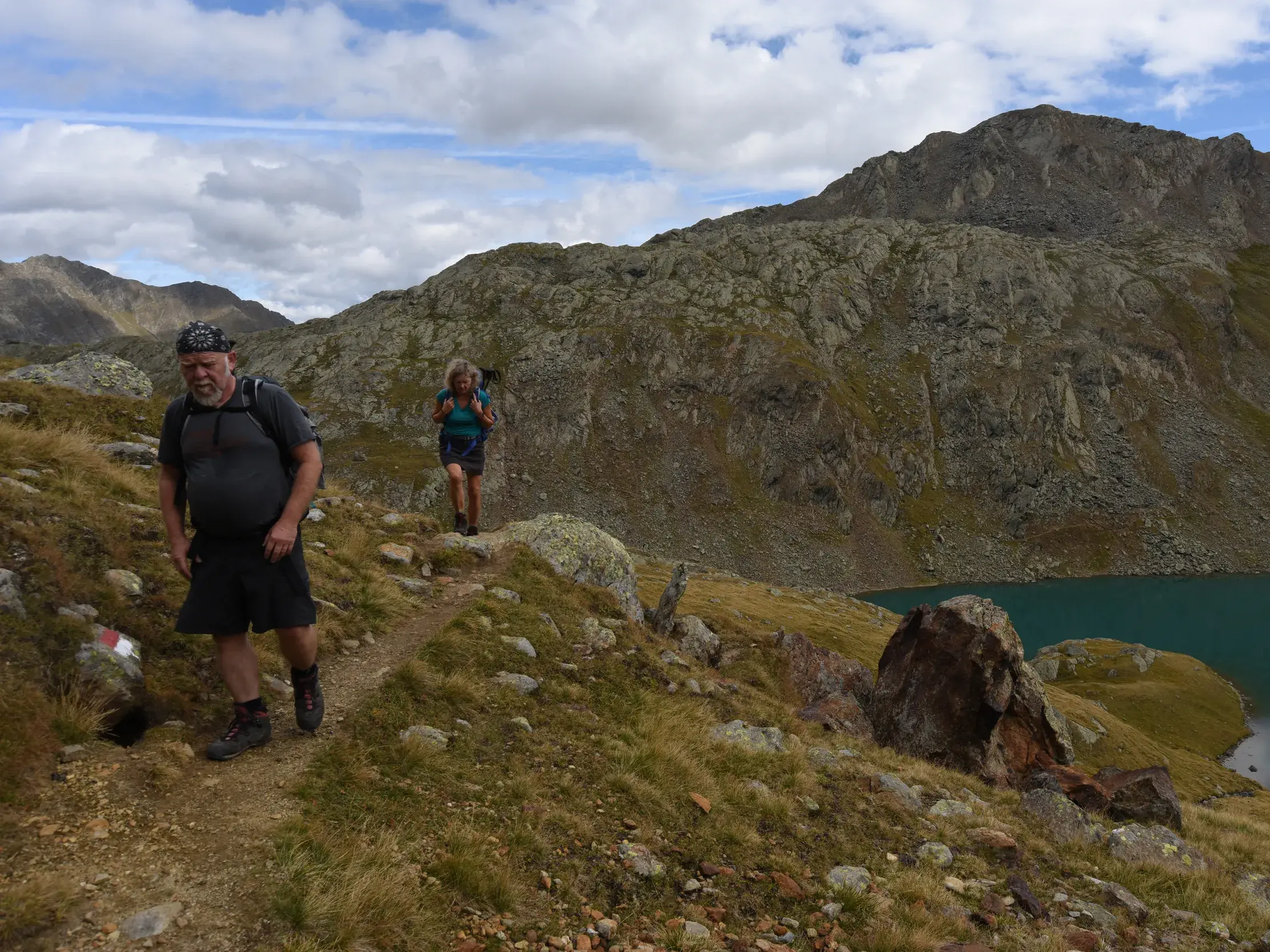 Zwei Wanderer mit Rucksäcken beim Trekking auf einem Bergpfad entlang eines türkisfarbenen Sees in den Stubaier Alpen.