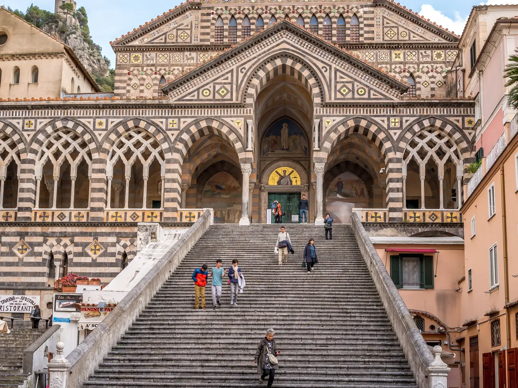 Besucher auf der monumentalen Freitreppe vor der reich verzierten, gestreiften Fassade des Doms Sant'Andrea in Amalfi.