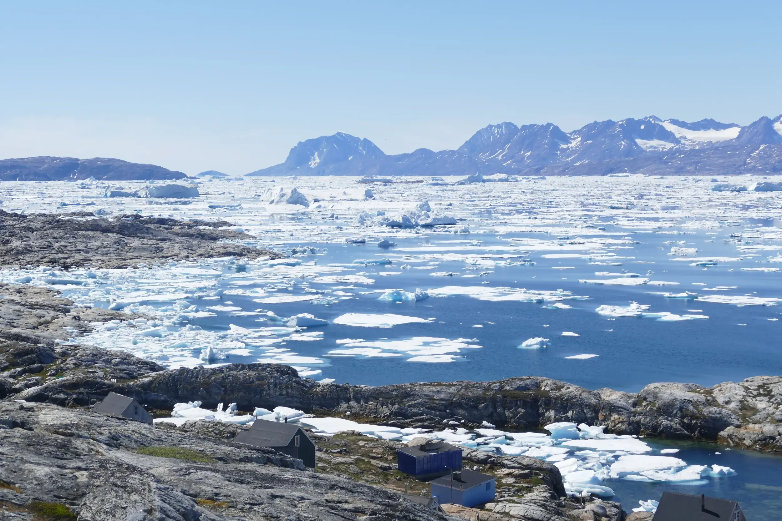 Holzhäuser der Inuit-Siedlung Tiniteqilaaq am Ufer des eisgefüllten Sermilik-Fjords mit Blick auf die Berge Ostgrönlands.