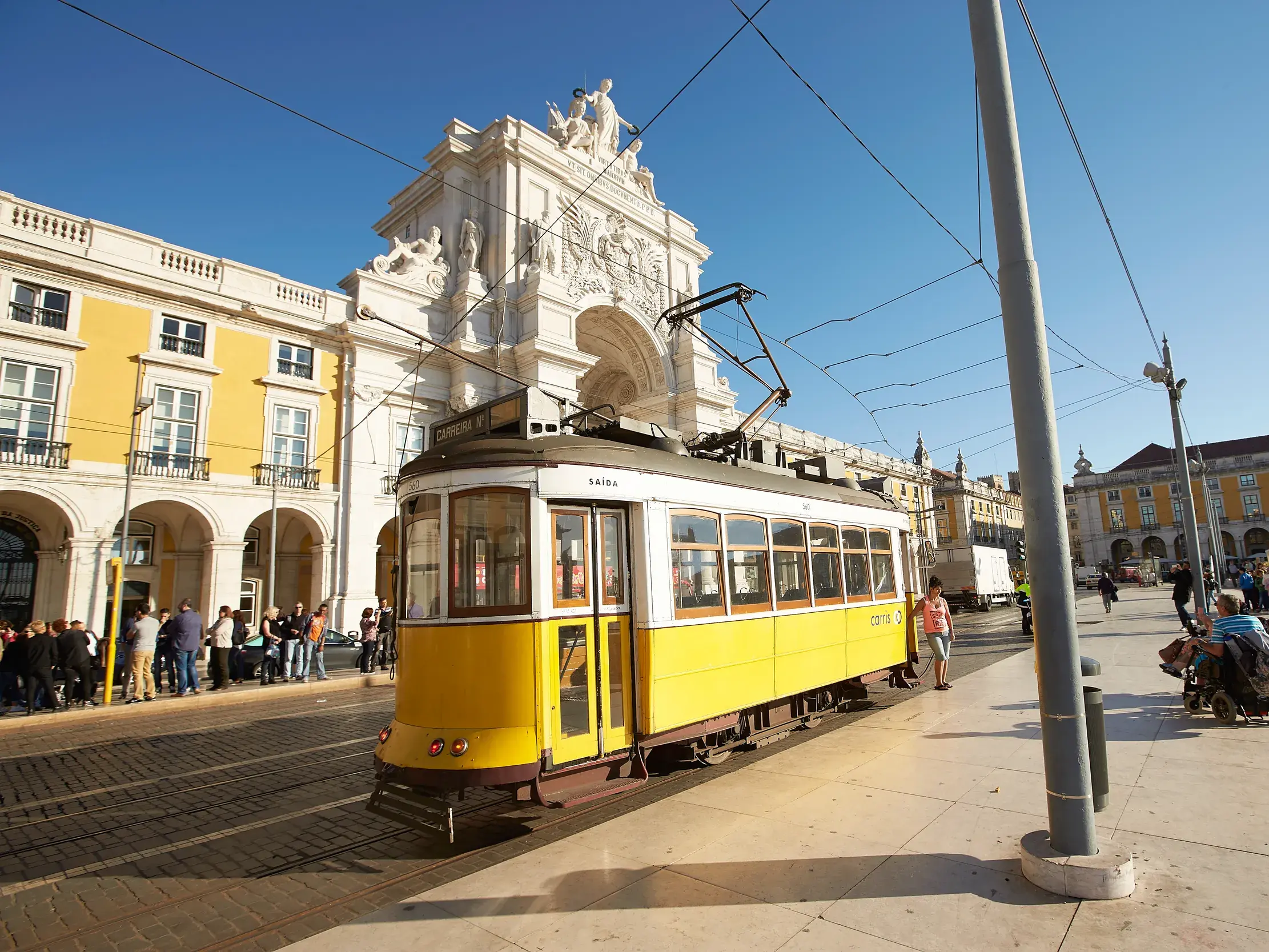 Eine gelbe Oldtimer-Straßenbahn hält an einem sonnigen Tag in Lissabon vor einem prachtvollen Triumphbogen und historischen Gebäuden.