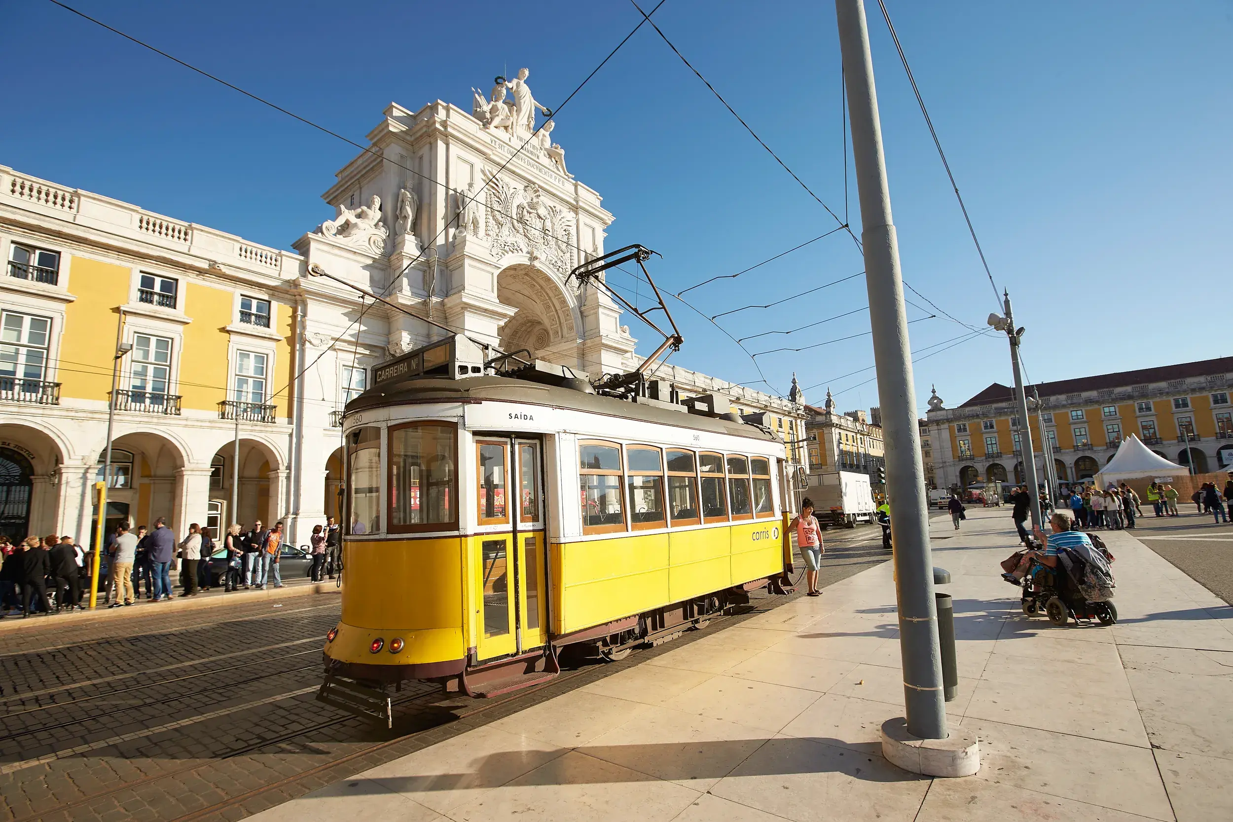 Eine gelbe Oldtimer-Straßenbahn hält an einem sonnigen Tag in Lissabon vor einem prachtvollen Triumphbogen und historischen Gebäuden.