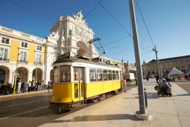 Eine gelbe Oldtimer-Straßenbahn hält an einem sonnigen Tag in Lissabon vor einem prachtvollen Triumphbogen und historischen Gebäuden.