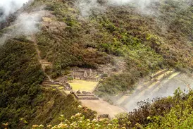 Blick von oben auf die nebelverhangenen Inka-Ruinen von Choquequirao, die in einen dicht bewachsenen Berghang in den Anden gebaut sind.
