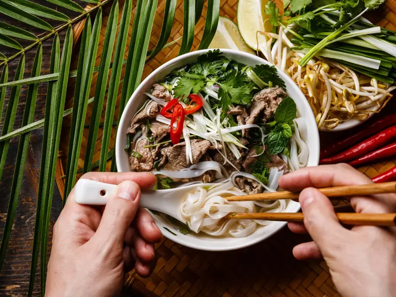 Hände mit Stäbchen und Löffel über einer Schale vietnamesischer Pho Bo als Stärkung auf einer Trekkingreise in Vietnam.
