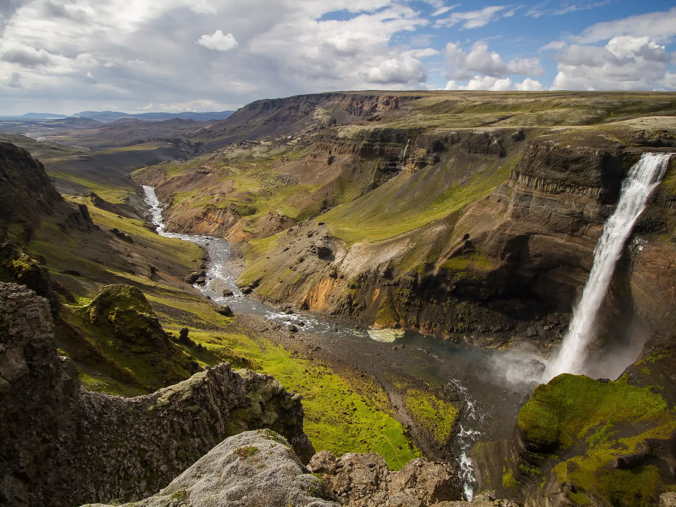 Der Wasserfall Haifoss in Island stürzt in eine tiefe, von moosgrünen Hängen gesäumte Schlucht, durch die ein Fluss fließt.