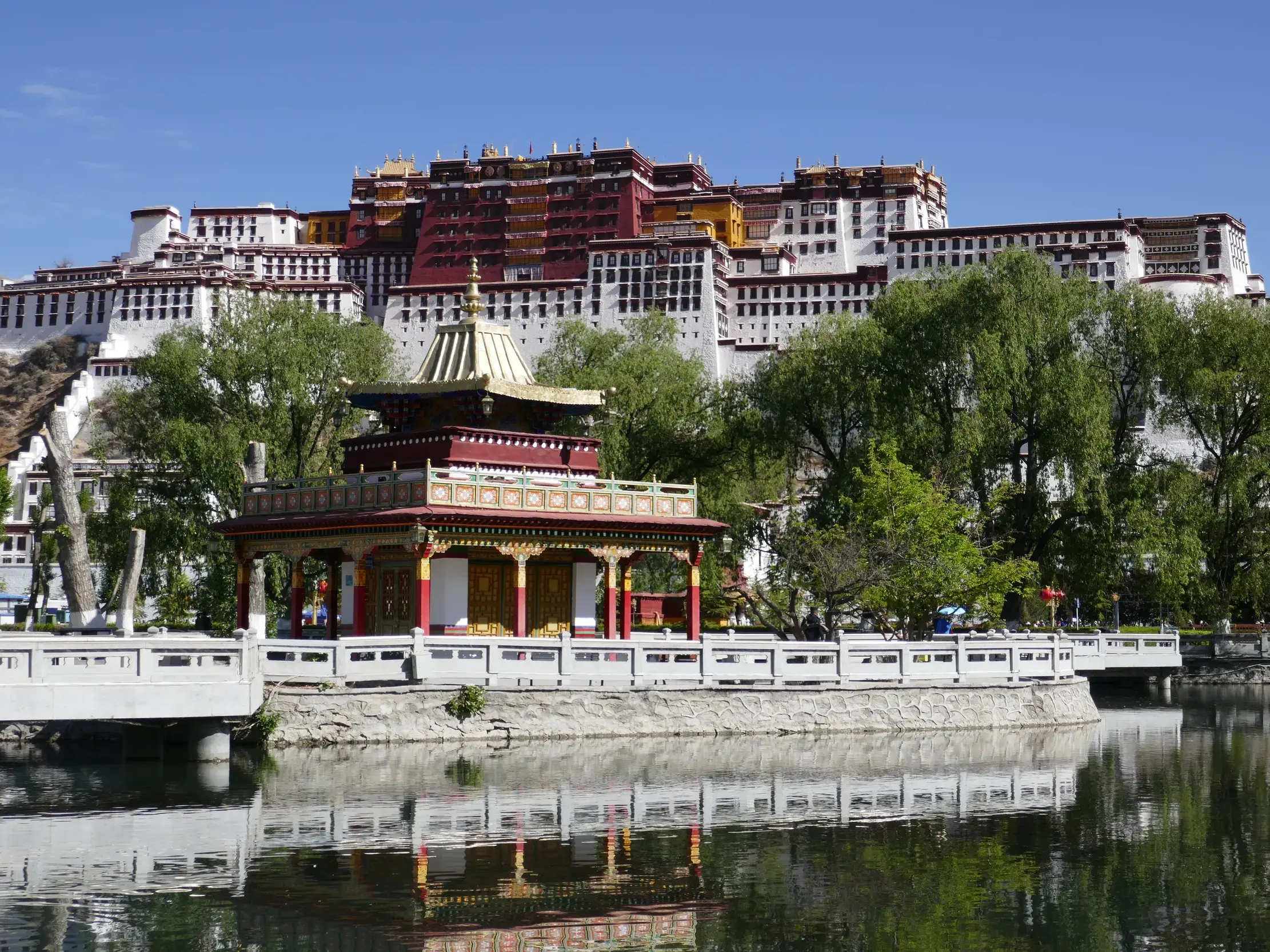 Blick über einen See mit traditionellem Pavillon auf den majestätischen Potala-Palast in Lhasa, Tibet, bei Sonnenschein.