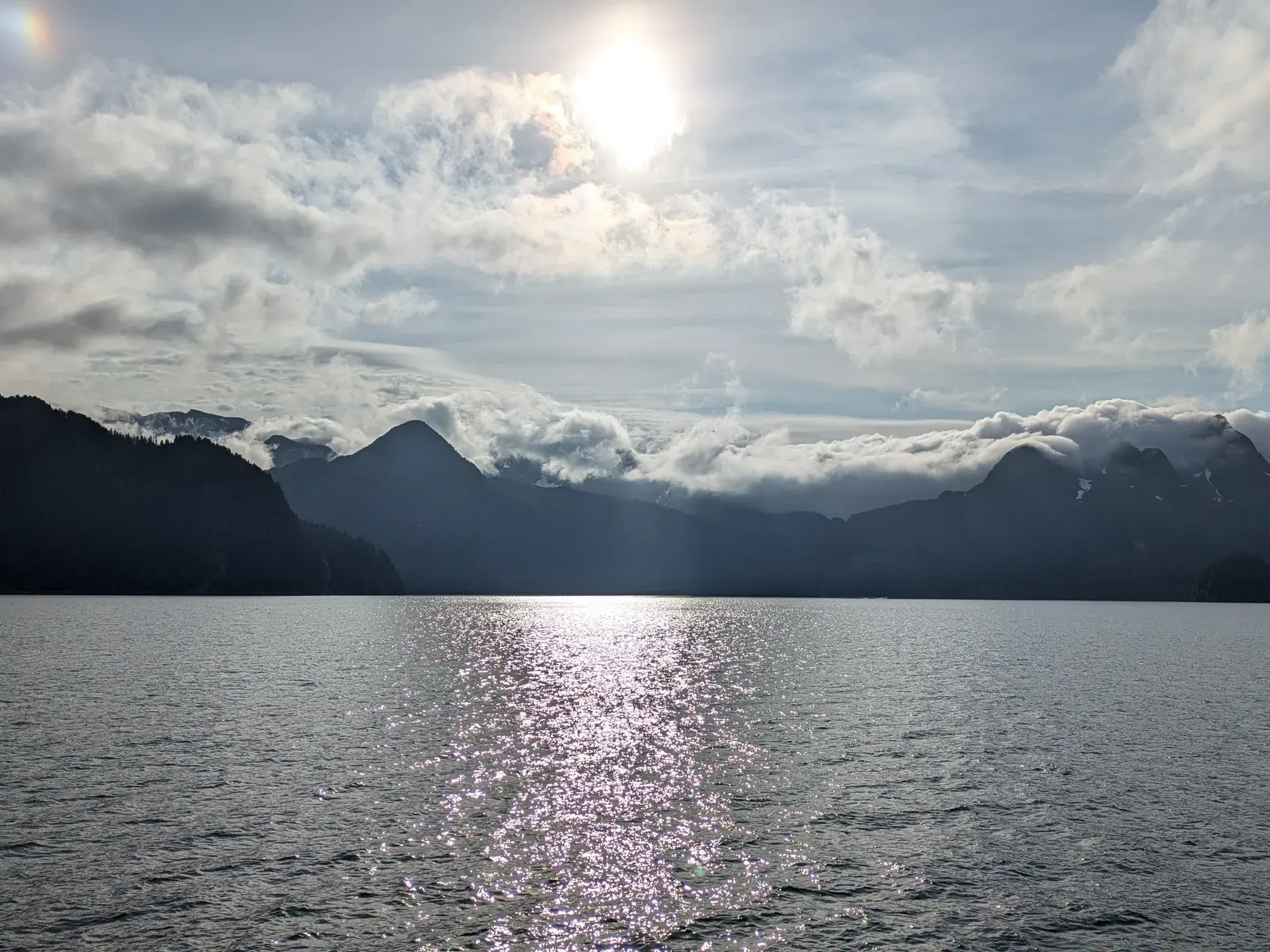 Die Sonne spiegelt sich auf dem Wasser des Kenai Fjords Nationalparks in Alaska vor der Silhouette wolkenverhangener Berge.