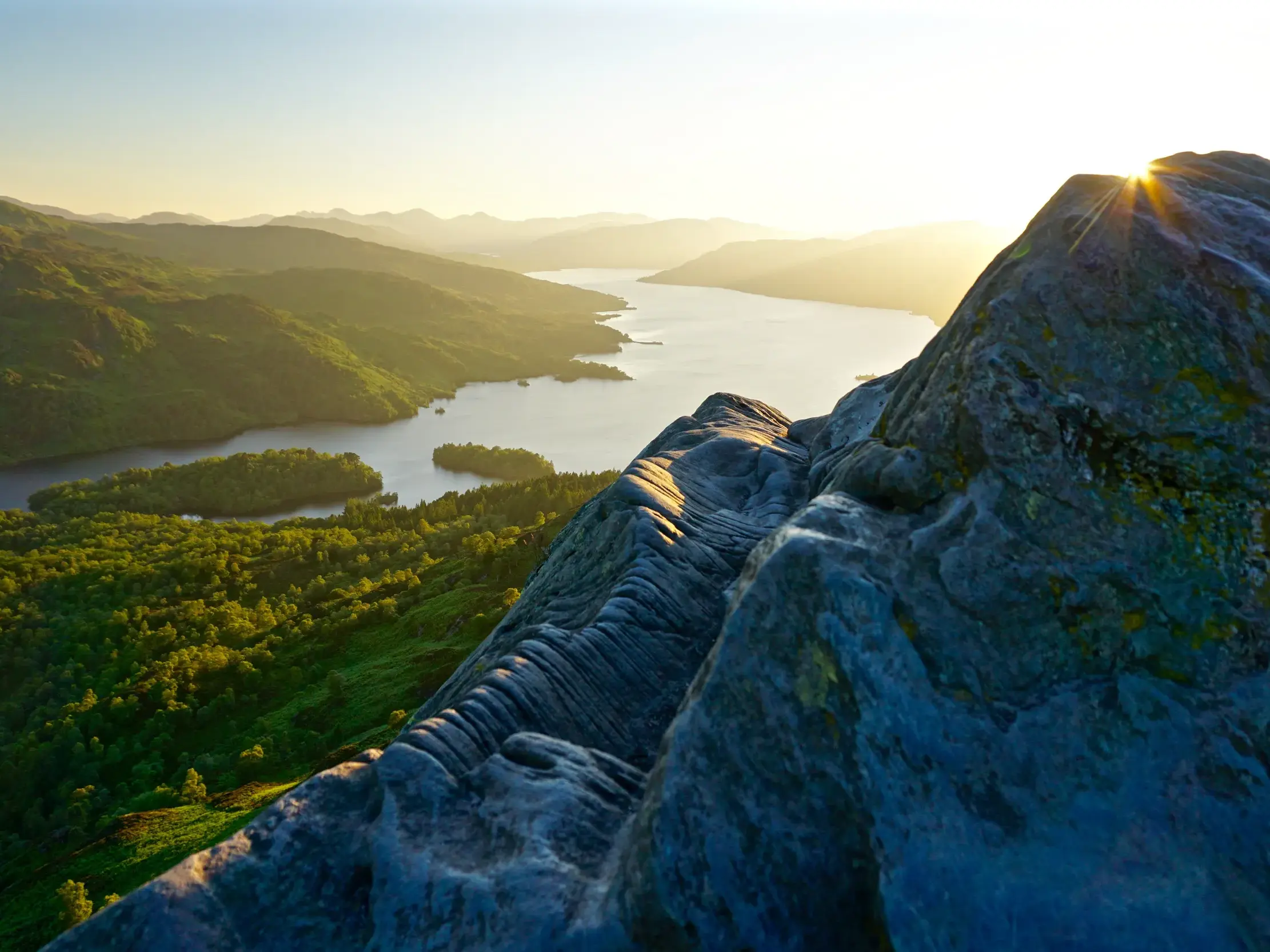 Felsiger Berggrat in den schottischen Highlands mit weitem Blick über den von Hügeln umgebenen Loch Katrine bei Sonnenaufgang.