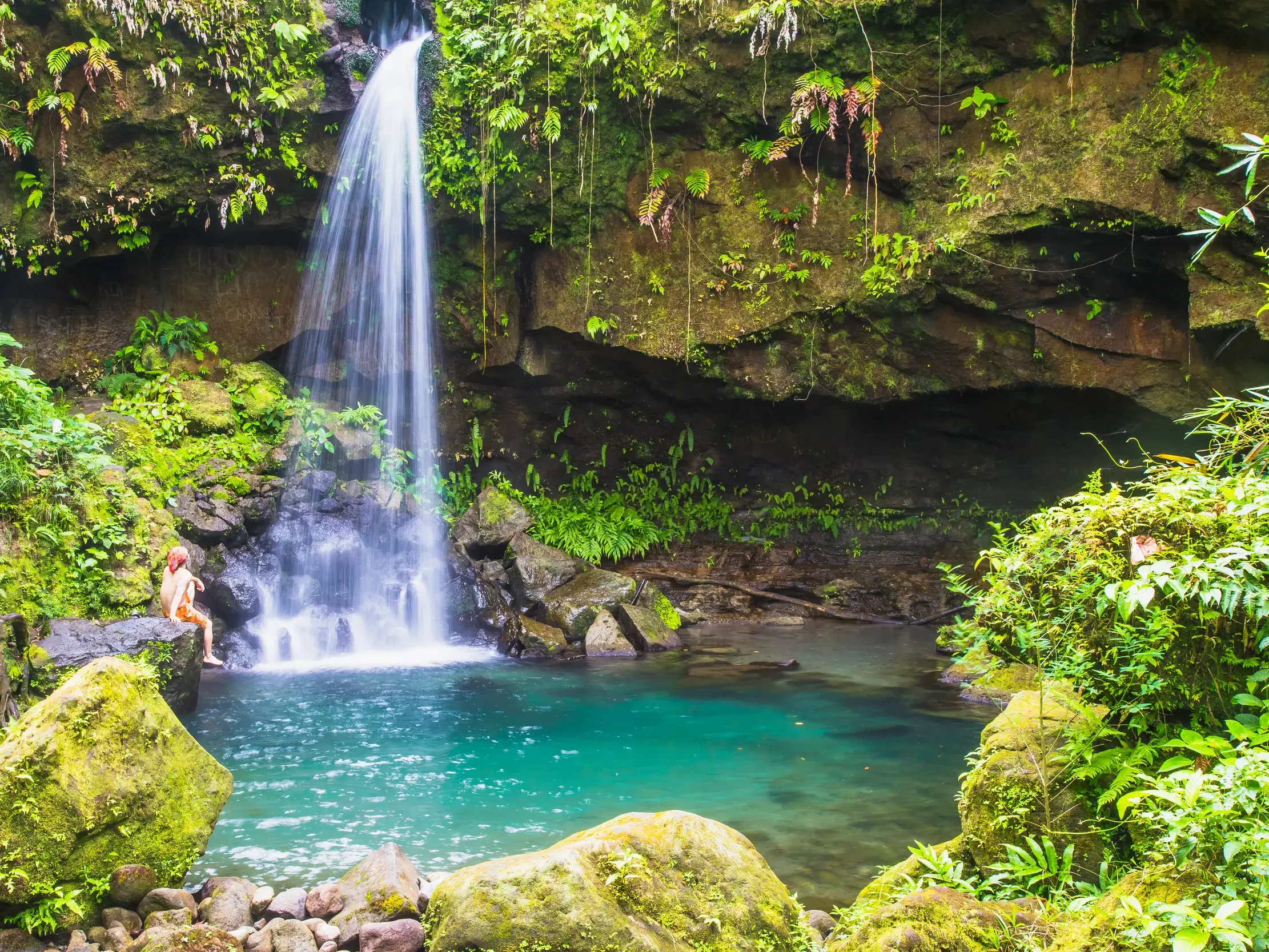 Person sitzt am Rande des Emerald Pools auf Dominica, während ein Wasserfall in das türkisfarbene Becken im Regenwald stürzt.