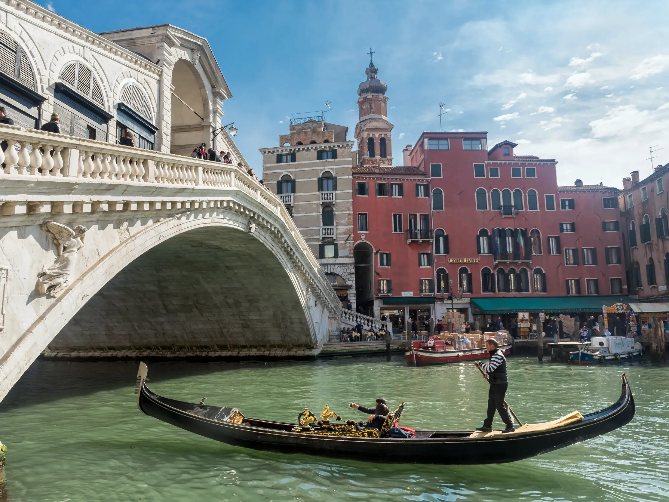 Eine Gondel gleitet auf dem Canal Grande in Venedig, daneben die steinerne Rialtobrücke und bunte Palazzi am Ufer.