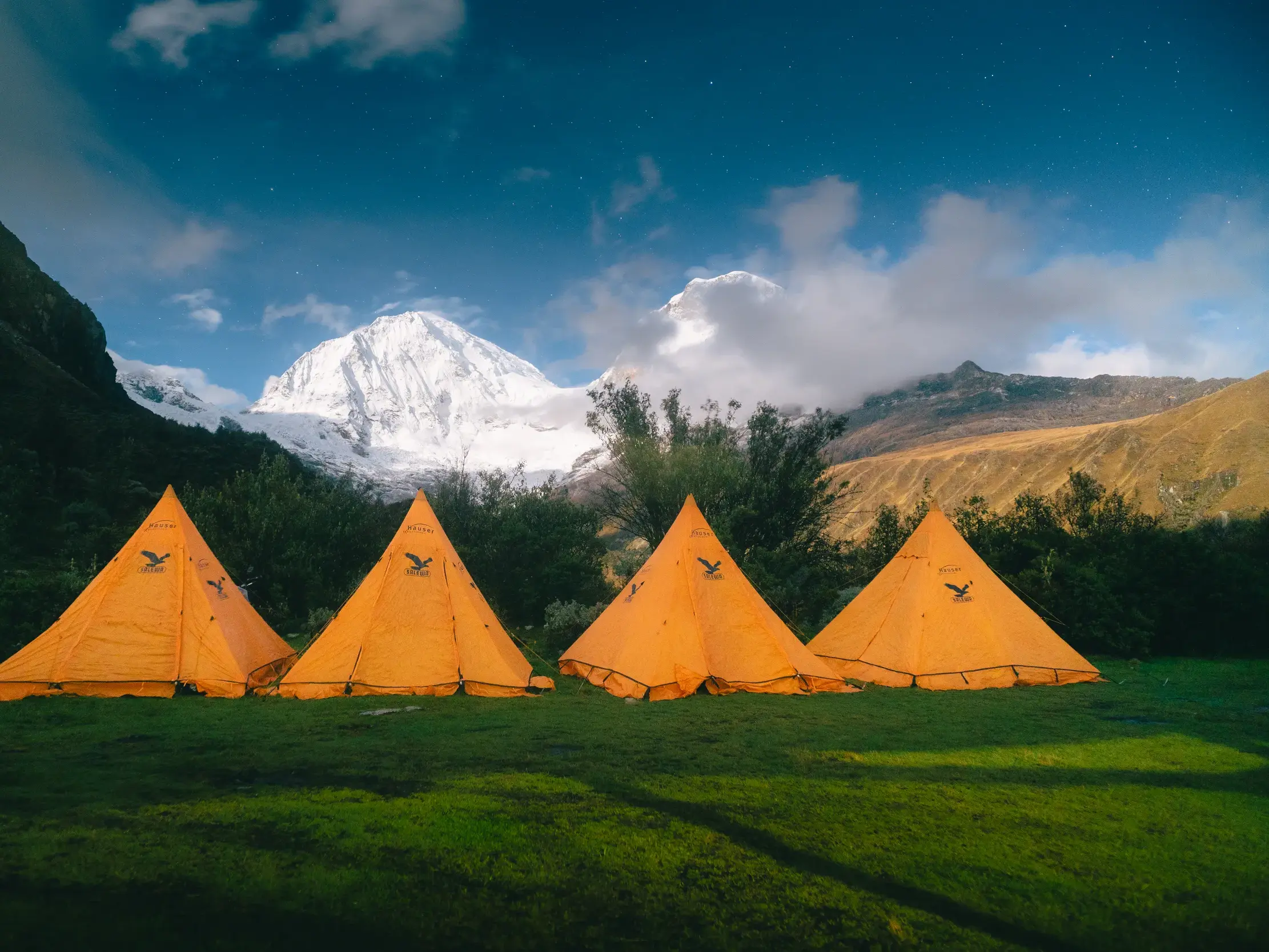 Vier orange Zelte auf einer grünen Wiese unter Sternenhimmel mit Blick auf einen schneebedeckten Gipfel in der Cordillera Blanca.