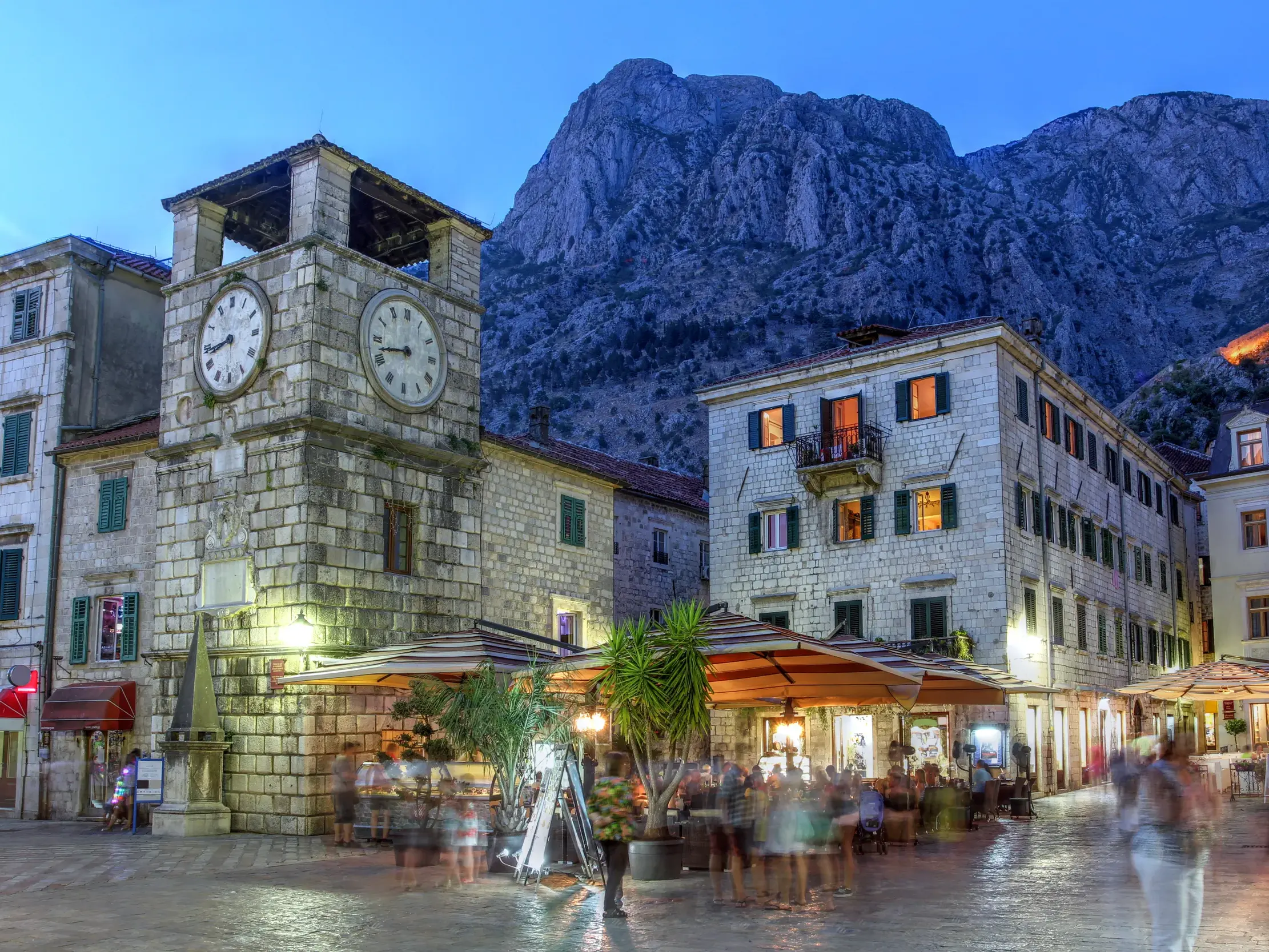 Der steinerne Uhrturm am belebten Waffenplatz in Kotor bei Dämmerung, im Hintergrund ragt ein Bergmassiv empor.