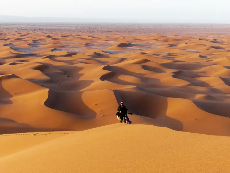 Ein Wanderer genießt die Aussicht von einer Sanddüne auf das orange leuchtende Dünenmeer des Erg Chegaga in der Sahara.