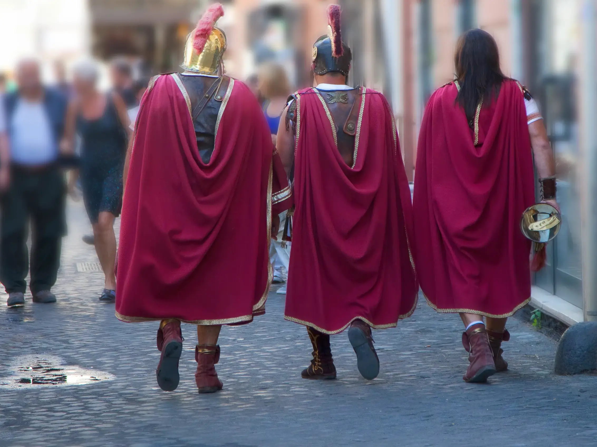 Drei als römische Legionäre verkleidete Darsteller mit roten Umhängen spazieren auf dem Kopfsteinpflaster in Rom.